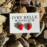 A pair of red strawberry stud earrings displayed on a white card labeled 'IVRY BELLE JEWELRY,' placed on a bed of small rocks outdoors.
