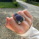 Hand wearing a silver ring with a tear drop shaped wampum set in a floral sterling silver ring outdoors on a gravel path.