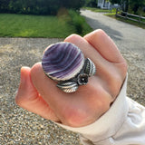 Hand wearing a ring with a circle shaped purple quahog shell with a flower and two leaves in sterling silver outdoors.