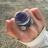 Hand wearing a ring with a purple and white quahog shell and silver band outdoors on a gravel path.