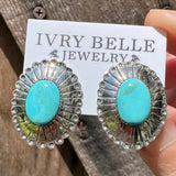 Turquoise stone earrings with silver setting on a wooden surface, accompanied by an 'Ivy Belle Jewelry' card.