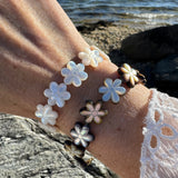 Close-up of a wrist wearing a bracelet with flower-shaped mother of pearl beads against a beach/ ocean background.