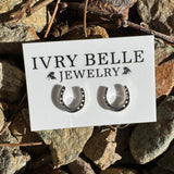A pair of small silver horseshoe stud earrings displayed on a white card labeled IVRY BELLE JEWELRY, placed on a bed of natural rocks outdoors.