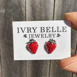 A hand holding a white jewelry card featuring a pair of strawberry stud earrings. The card is branded 'IVRY BELLE JEWELRY' and is photographed against a rustic gray wooden plank background.