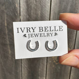 A hand holding a white display card with two small silver horseshoe hoop earrings. The card is branded 'IVRY BELLE JEWELRY' and is photographed against a rustic gray wooden plank background.