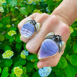 Hand wearing two purple and white wampum stone rings with gold bands against a green floral background