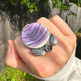 Hand wearing a large ring with a round purple and white quahog shell with floral accents outdoors.