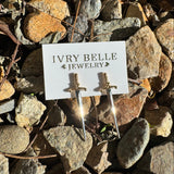 A pair of gold-tone dagger stud earrings displayed on a white card labeled 'IVRY BELLE JEWELRY,' placed on a bed of small brown and gray rocks outdoors.
