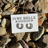 A pair of small silver hoop earrings displayed on a white card labeled 'IVRY BELLE JEWELRY,' placed on a bed of gray and brown rocks outdoors.