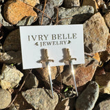 A pair of small dagger-shaped earrings displayed on a white card labeled 'IVRY BELLE JEWELRY,' placed on a bed of natural rocks outdoors.