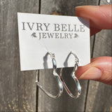 A pair of silver hoop earrings displayed on a white card labeled 'IVRY BELLE JEWELRY', held by a hand against a weathered wooden plank background.