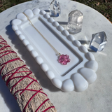 White ceramic jewelry dish with a necklace and pink flower pendant, surrounded by crystals on a marble surface.