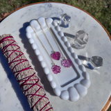 Jewelry dish with pink heart-shaped earrings, crystals, and a smudge stick on a marble surface.