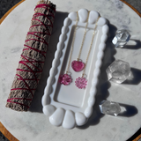 Necklace with pink heart pendants on a white stand, next to a smudge stick and crystals on a marble surface.