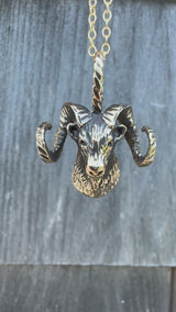 A bronze ram head necklace dangling in front of a wooden background.