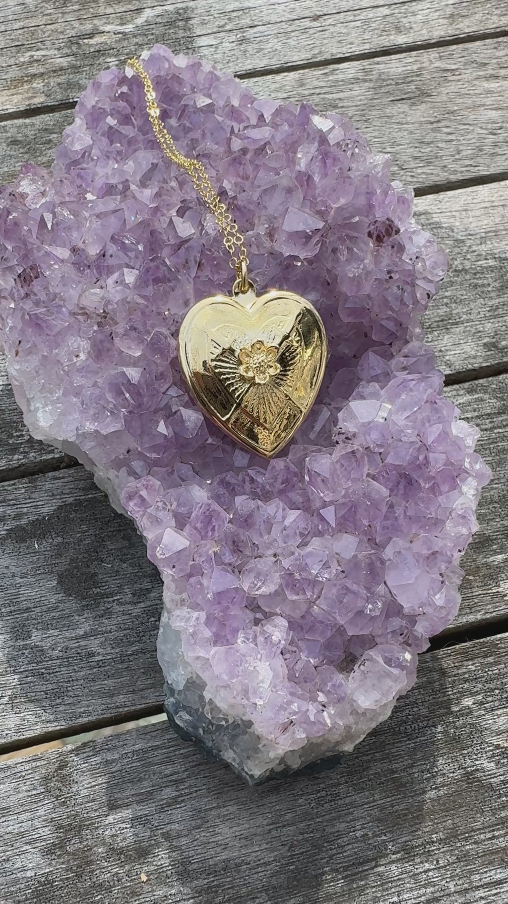A video of a gold heart necklace resting upon a purple amethyst crystal on a gray wooden background.