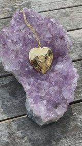 A video of a gold heart necklace resting upon a purple amethyst crystal on a gray wooden background.