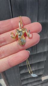 A gold ram head necklace held in a hand against a wooden background