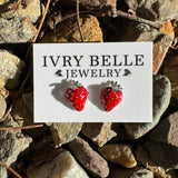 A pair of strawberry-shaped stud earrings in red with white speckles and silver tops, displayed on a white card labeled 'IVRY BELLE JEWELRY' placed on a bed of rocks outdoors